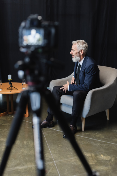uomo d'affari barbuto in giacca e cravatta seduto in poltrona durante l'intervista vicino alla fotocamera digitale offuscata su treppiede - Foto, immagini