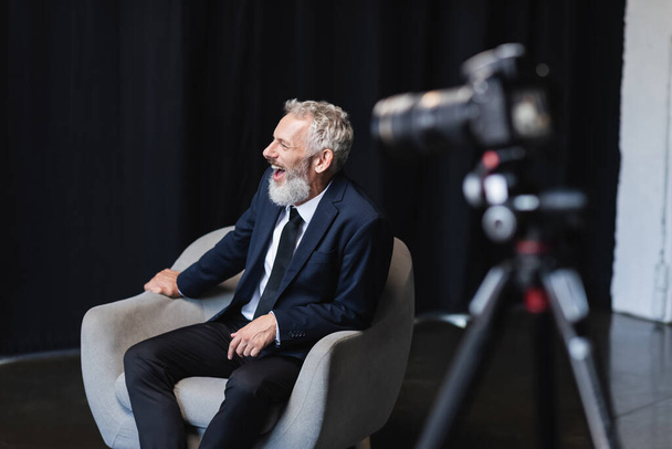 alegre hombre de negocios en traje riendo mientras se sienta en sillón durante la entrevista cerca de la cámara digital borrosa en el trípode - Foto, Imagen