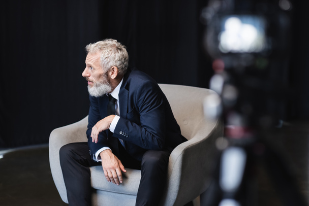 businessman in suit sitting in grey armchair during interview near blurred digital camera in studio  - Photo, Image