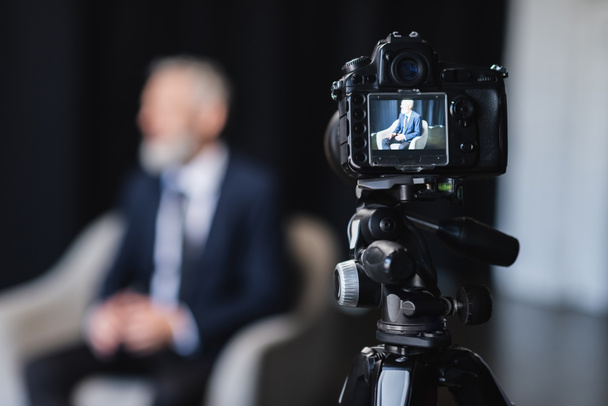 ψηφιακή φωτογραφική μηχανή με επιχειρηματία με κοστούμι κάθεται σε γκρι πολυθρόνα κατά τη διάρκεια της συνέντευξης στην οθόνη - Φωτογραφία, εικόνα