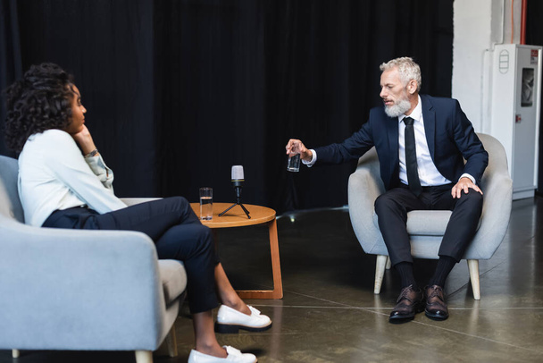 uomo d'affari in giacca e cravatta che prende un bicchiere d'acqua durante un'intervista presso il giornalista afroamericano  - Foto, immagini