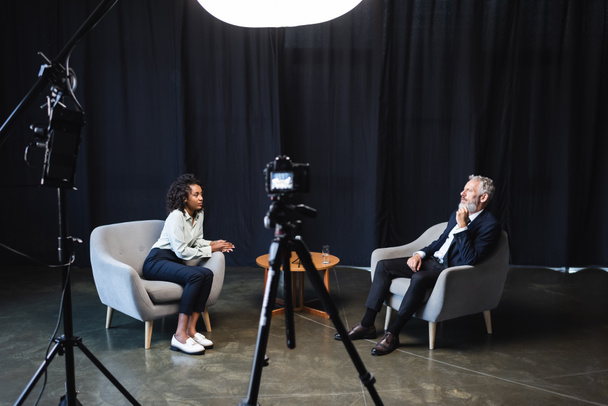 хвилястий афро-американський журналіст сидить у кріслі і розмовляє з гостем у студії  - Фото, зображення