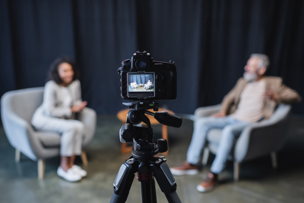 cámara digital con hombre de negocios en traje sentado en sillón gris durante entrevista con periodista afroamericano en pantalla  - Foto, Imagen