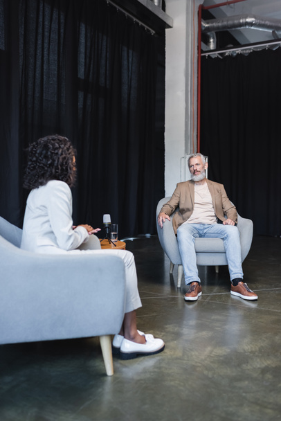 kudrnatý africký americký novinář v obleku mluví s vousatým podnikatelem sedí v křesle během rozhovoru - Fotografie, Obrázek