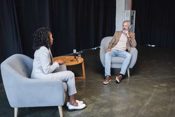 kudrnatý africký americký novinář v obleku mluví s podnikatelem sedí v křesle během rozhovoru - Fotografie, Obrázek