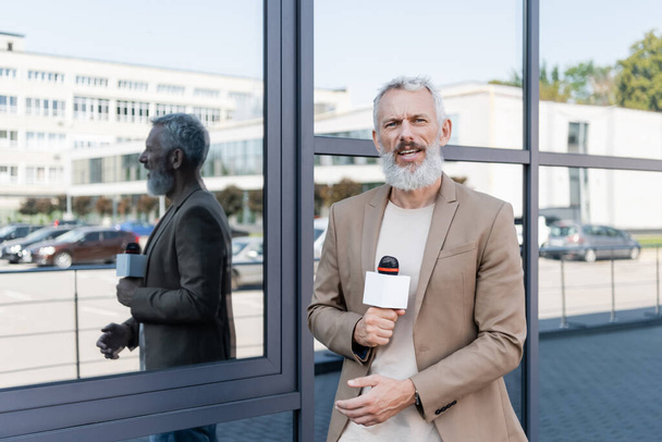 szakállas riporter blézerben, mikrofonnal a kezében, és riportot készít az épület közelében.  - Fotó, kép