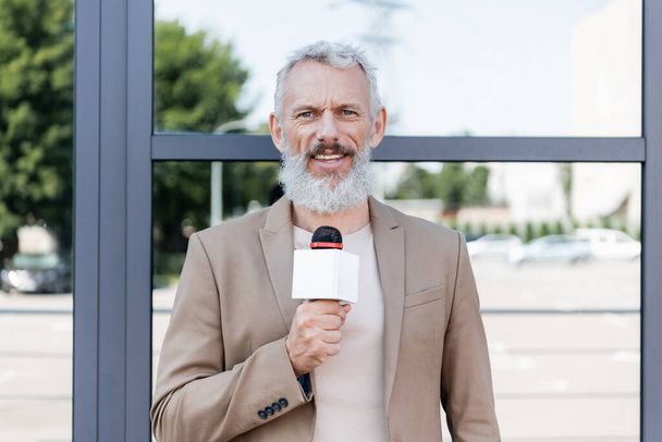 bearded announcer in blazer holding microphone and making reportage near building  - Φωτογραφία, εικόνα