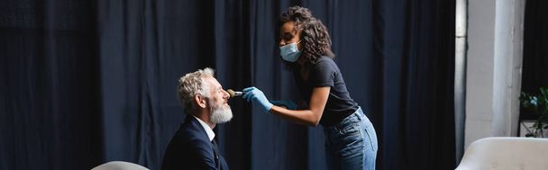 krullend Afrikaans amerikaans make-up kunstenaar in medisch masker en latex handschoenen aanbrengen gezicht poeder op gezicht van zakenman, banner - Foto, afbeelding