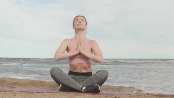 Stab shot van jonge fitte vrouw in sportkleding zittend op yoga mat in lotus poseren mediteren met biddende handen tijdens outdoor yoga praktijk op het strand in de ochtend - Video