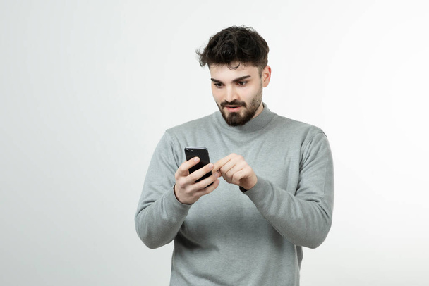 Portrait d'un gars attrayant tenant un téléphone intelligent contre un mur blanc. Photo de haute qualité - Photo, image