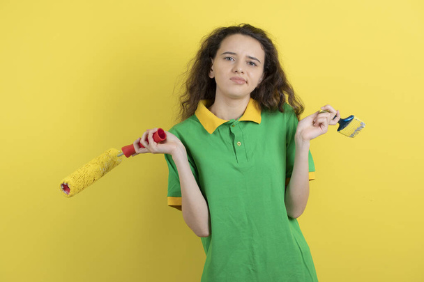 Fiatal tini lány kezében festék tekercs és ecset sárga háttér. Kiváló minőségű fénykép - Fotó, kép