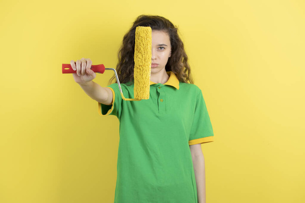 Joven adolescente sosteniendo rollo de pintura y de pie sobre fondo amarillo. Foto de alta calidad - Foto, Imagen
