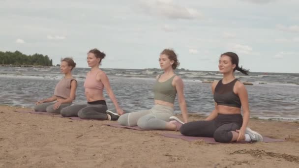 Langzame opname van vier jonge vrouwen die samen strandyoga beoefenen, zittend op yogamatten naast helder blauw water op winderige ochtend - Video