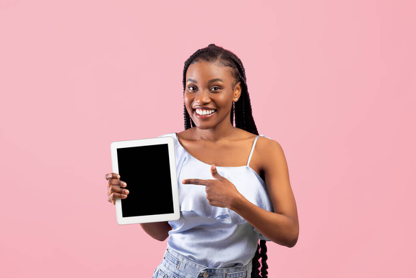 Positive young Afro woman pointing at tablet computer with empty screen on pink background, mockup for website or app - Foto, Imagem