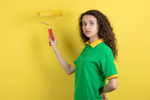 Young teen girl holding paint roll and standing on yellow background. High quality photo - Photo, Image