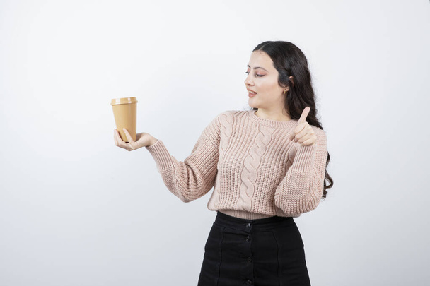 Giovane donna bruna mostrando pollice in su e tenendo una tazza di caffè caldo. Foto di alta qualità - Foto, immagini