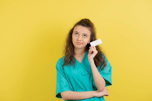Молода медсестра в уніформі дивиться на медичні таблетки над жовтою стіною. Високоякісна фотографія
 - Фото, зображення
