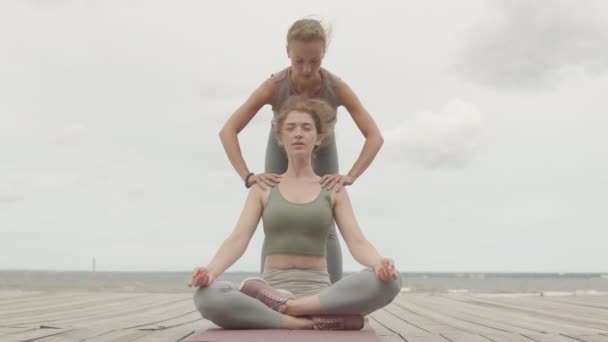 Медленный снимок молодой белой женщины, сидящей в позе лотоса, практикующей йогу на открытом воздухе на пирсе в ветреное утро с женщиной-тренером, помогающей ей - Кадры, видео