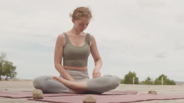 Slowmo plan de jeune femme concentrée en pose de lotus méditant seul assis sur un tapis de yoga à l'extérieur sur une jetée en bois par une journée nuageuse et venteuse - Séquence, vidéo