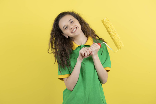 Imagen de chica rizada en camiseta verde posando con rodillo de pintura sobre fondo amarillo. Foto de alta calidad - Foto, Imagen