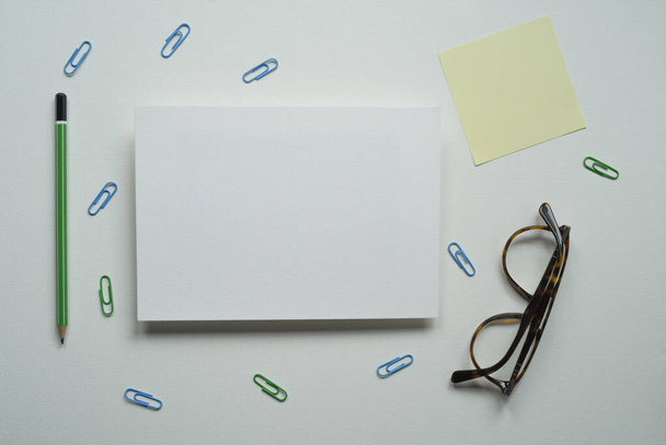 escritorio, tarjeta blanca, rectángulo, espacio para texto, diseño, lápiz verde, recordatorio, tarjeta amarilla, gafas marrones, clips de papel, sobre un fondo blanco, foto de arriba - Foto, Imagen
