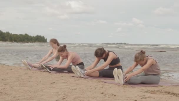 Slowmo shot of for young Caucasian women in sportswear doing stretching exercises on yoga mats, sitting in row on sandy beach on windy morning - Footage, Video