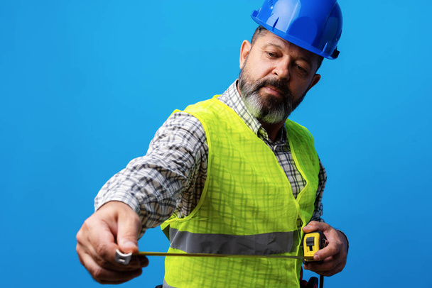 Male builder holding measuring roulette against blue background - Foto, afbeelding