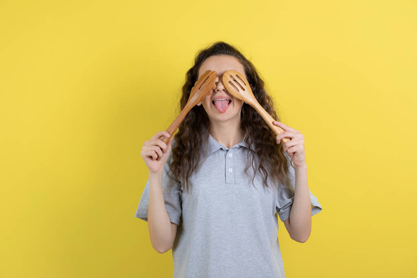 Beautiful young girl covering her eyes with kitchen tools standing on yellow background. High quality photo - Фото, изображение