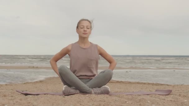 Colpo di pugnalata sorridente giovane donna caucasica in abbigliamento sportivo meditando da solo sulla spiaggia sabbiosa il mattino ventoso e nuvoloso - Filmati, video