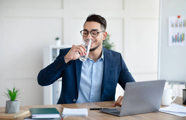Крутой арабский бизнесмен в костюме питьевой воды во время работы на ноутбуке в современном офисе - Фото, изображение