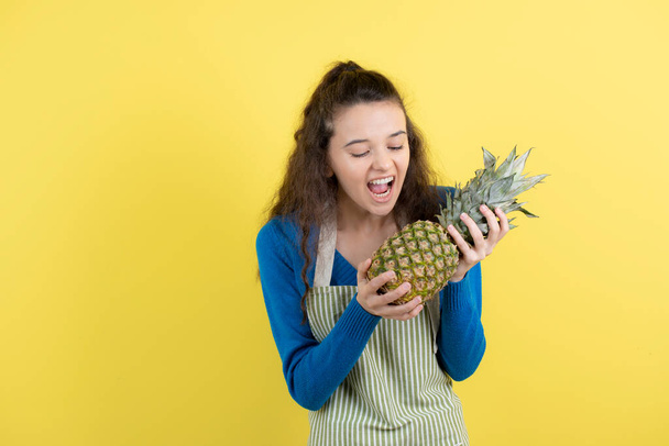 Young curly teen in apron trying to eat ripe pineapple over yellow wall. High quality photo - Photo, Image