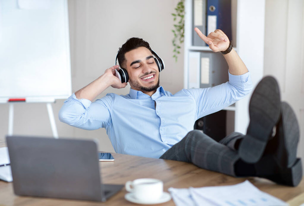 Перерыв в офисе. Радостный араб слушает музыку в беспроводных наушниках, расслабляется на рабочем месте - Фото, изображение