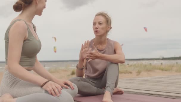 Powolne ujęcie młodej kobiety rozmawiającej z trenerką jogi siedzącej razem na drewnianym pomoście podczas treningu jogi na świeżym powietrzu w pochmurny i wietrzny dzień - Materiał filmowy, wideo