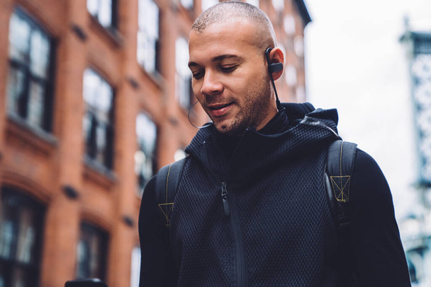 Thoughtful bold bristly athletic man in headphones with rucksack and in black sportswear using mobile in daylight on urban blurred background - Fotó, kép
