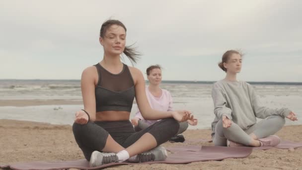 Slowmo tiro de grupo de jovens mulheres caucasianas sentadas em lótus posar na praia de areia na manhã ventosa e nublada meditando com os olhos fechados - Filmagem, Vídeo