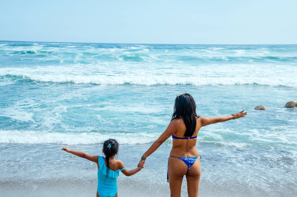 Πορτραίτο δράσης της νεαρής μητέρας με την κόρη στην πλάτη της μπροστά στη θάλασσα. Κορίτσια με μαγιό σηκώνουν τα χέρια. - Φωτογραφία, εικόνα