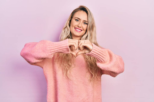 Красива молода блондинка в рожевому светрі посміхається, показуючи серцевий символ і форму руками. романтична концепція
.  - Фото, зображення