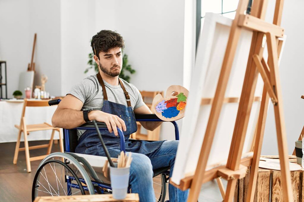 Jeune homme hispanique assis sur la peinture en fauteuil roulant au studio d'art déprimé et inquiet pour la détresse, pleurer en colère et peur. triste expression.  - Photo, image