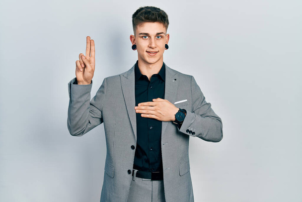 Jeune garçon caucasien avec dilatation des oreilles portant une veste d'affaires souriant jurant avec la main sur la poitrine et les doigts vers le haut, faisant un serment de fidélité  - Photo, image