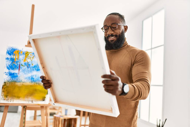 Νεαρός Αφροαμερικάνος καλλιτέχνης χαμογελά χαρούμενος κρατώντας καμβά στο στούντιο τέχνης. - Φωτογραφία, εικόνα