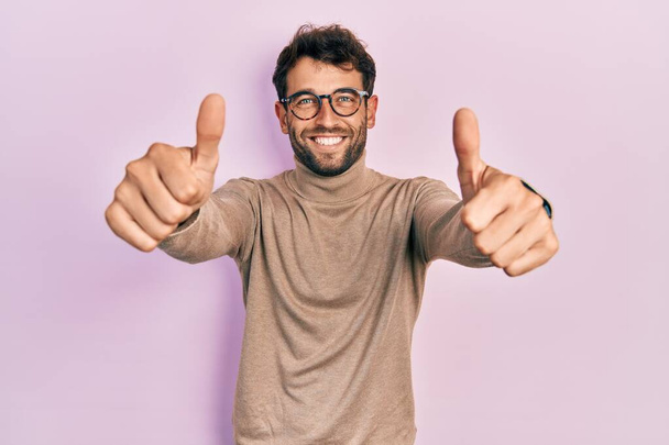 Handsome man with beard wearing turtleneck sweater and glasses approving doing positive gesture with hand, thumbs up smiling and happy for success. winner gesture.  - Photo, Image
