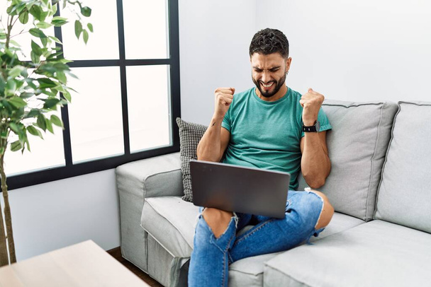 Young handsome man with beard using computer laptop sitting on the sofa at home excited for success with arms raised and eyes closed celebrating victory smiling. winner concept.  - Photo, Image