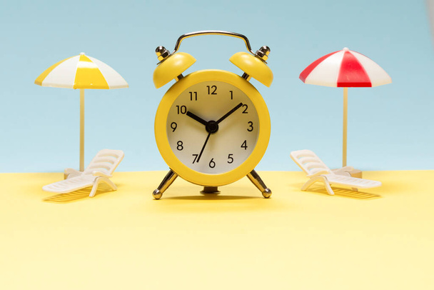 旅行の時間です。黄色、青の背景にサンラウンジャー、黄色の傘と目覚まし時計. - 写真・画像