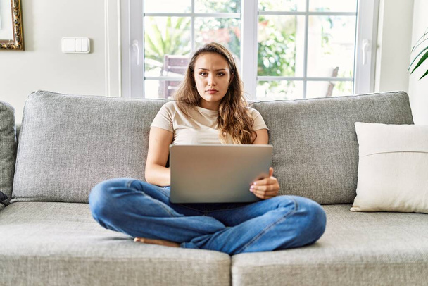 自宅でコンピュータのラップトップを使用してソファの上に座っている美しい若いブルネットの女性は、顔に深刻な表情でリラックスしました。カメラを見ているだけで.  - 写真・画像