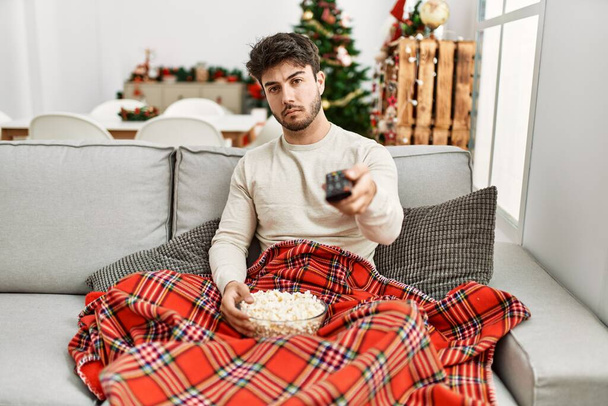 Mladý hispánec jíst popcorn sedí na pohovce u vánočního stromku myšlení postoj a střízlivý výraz hledá sebevědomí  - Fotografie, Obrázek