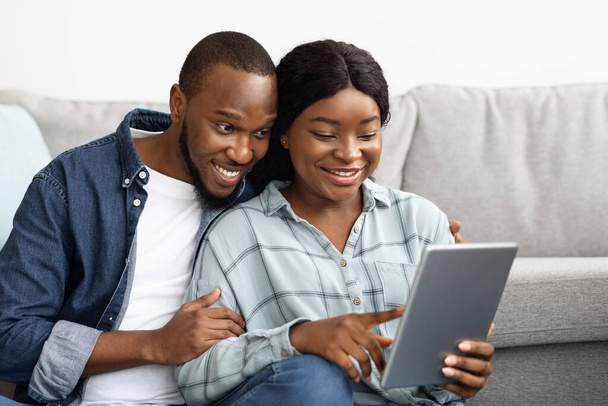 Μαύρο ευτυχισμένο Millennial ζευγάρι χρησιμοποιώντας ψηφιακή ταμπλέτα μαζί, ενώ χαλαρώνοντας στο σπίτι - Φωτογραφία, εικόνα