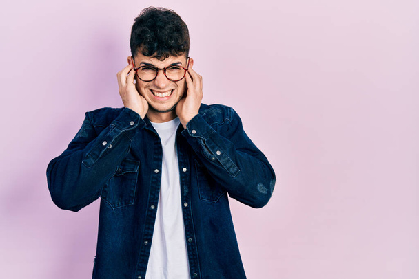 Молодий іспаномовний чоловік носить повсякденний одяг і окуляри, прикриваючи вуха пальцями з роздратованим виразом за шум гучної музики. глуха концепція
.  - Фото, зображення