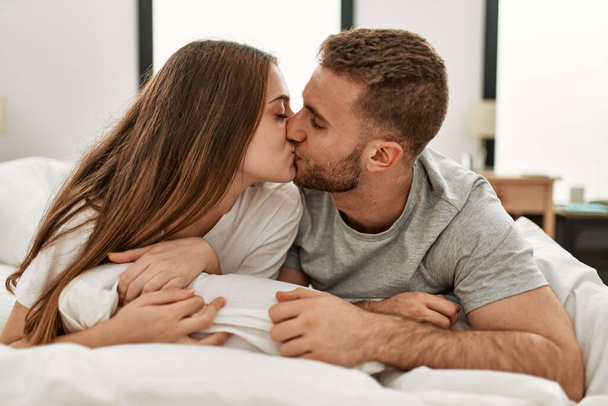 Νεαρό καυκάσιο ζευγάρι χαμογελά ευτυχισμένη και φιλιά στο κρεβάτι στο σπίτι. - Φωτογραφία, εικόνα