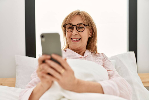 Moyen-âge femme blonde en utilisant smartphone couché sur le lit à la maison. - Photo, image