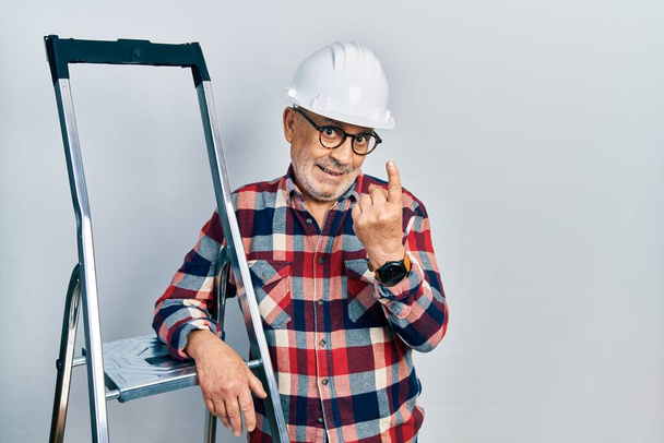 Handyman maduro guapo cerca de escaleras de construcción usando hardhat señas vienen aquí gesto con la mano invitando a dar la bienvenida feliz y sonriente  - Foto, imagen